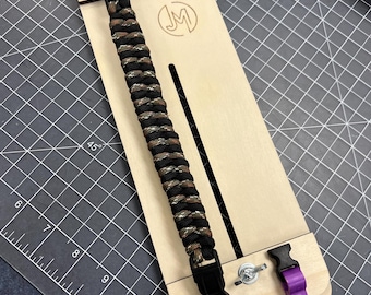 May include: A wooden paracord jig with a black and brown paracord bracelet being made on it. The jig has purple clips and a metal screw. The jig has the letters "JM" engraved on it.
