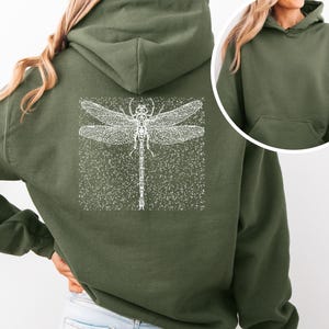 Può includere: Una felpa con cappuccio verde con una grafica di libellula bianca sul retro. La felpa ha una vestibilità rilassata e un cappuccio con coulisse.