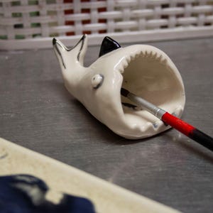 Puede incluir: Un soporte para pinceles con forma de tiburón de cerámica blanca, con detalles en negro y plateado. La boca del tiburón sostiene un pincel con mango rojo y negro. El soporte está sobre una superficie gris.