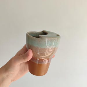 Könnte beinhalten: Eine handgefertigte Keramik-Tasse mit einer einzigartigen Form und einem zweifarbigen Farbschema. Der obere Bereich ist helltürkis, die Mitte ist warmbraun und der Boden ist dunkler braun. Die Tasse hat ein unverwechselbares Griffdesign.