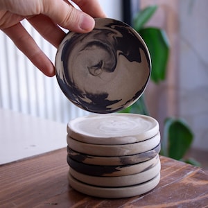May include: A set of handmade ceramic coasters. Each coaster is round, with a beige base and a distinctive swirling black pattern. One coaster is held above the stack, displaying the design. The coasters are on a wooden surface.