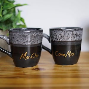 May include: Two black ceramic mugs with a speckled gray rim and gold lettering. One mug reads "Mon Cher" and the other reads "Cara Mia". The mugs have a curved handle and sit on a wooden surface.