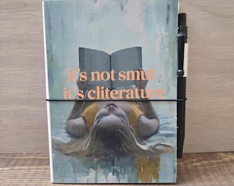 May include: A notebook with the text "it's not smut it's cliterature" in orange lettering. The cover features an illustration of a person reading a book. A black pen is attached to the notebook with a black elastic band.