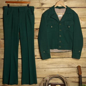 Könnte beinhalten: Ein dunkelgrünes zweiteiliges Set bestehend aus einer langärmeligen Jacke und einer passenden Hose. Die Jacke hat einen Kragen, eine Knopfleiste, zwei Brusttaschen mit Klappen und geknöpfte Manschetten. Die Hose hat einen geraden Schnitt.