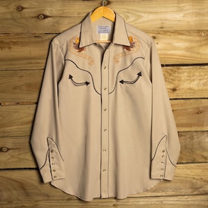 Op de afbeelding: Een beige overhemd met lange mouwen en knoopsluiting, voorzien van donkerbruine biezen langs het voorpand, de kraag en de manchetten. Het overhemd heeft geborduurde adelaarsmotieven op de borst en een label met H Bar C California Ranchwear.