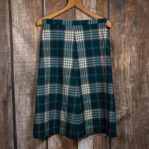 May include: A vintage green plaid skirt hanging on a wooden hanger. The skirt features a classic plaid pattern in shades of green, white, and red. The skirt is knee-length and appears to be made of a wool or wool-blend fabric.