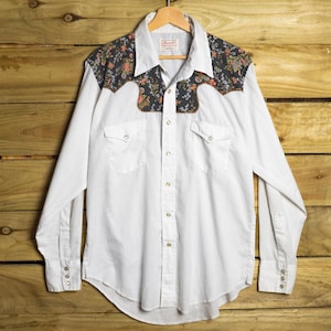 May include: A long-sleeve button-down shirt featuring a solid white body and a dark grey floral patterned yoke across the shoulders. The shirt includes two chest pockets with flaps, a pointed collar, and metallic buttons down the front placket.