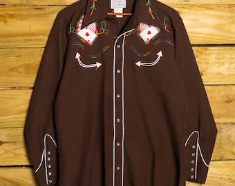 May include: A brown long-sleeve shirt with white piping and decorative embroidery. The shirt features playing card designs and white arrow accents. The cuffs have button closures. The shirt is hanging on a wooden hanger.