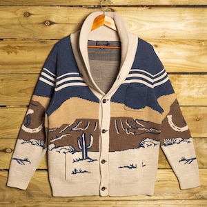 Könnte beinhalten: Beige, blau und brauner Cardigan-Pullover mit Wüstenlandschaftsdesign. Der Pullover hat einen Schalkragen, einen Knopfverschluss und eine Tasche. Das Design umfasst einen Kaktus, Berge und Streifen.