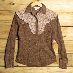Op de afbeelding: Een bruin shirt met lange mouwen met een bloemenpatroon en een geplooid juk. Het juk is crèmekleurig met een klein bloemmotief. Het shirt heeft een knoopsluiting en manchetten en hangt aan een houten hanger.