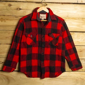 Könnte beinhalten: Rot-schwarz kariertes Flanellhemd mit zwei Brusttaschen und Kragen. Das Hemd hängt an einem Holzbügel vor einem Holzgrund. Die Marke "Woolrich" ist auf einem Etikett zu sehen.