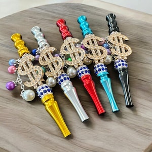 May include: An assortment of five decorative smoking accessories in various colors, including gold, silver, red, blue, and black. Each features a rhinestone-studded dollar sign charm and a beaded accent. The accessories are arranged on a wooden surface.