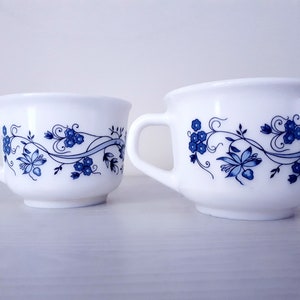 May include: Two white ceramic mugs of different sizes featuring a blue floral pattern. Each mug has a single handle and a wide rim. The design consists of blue flowers, swirling vines, and leaves wrapped around the exterior of the white cups.