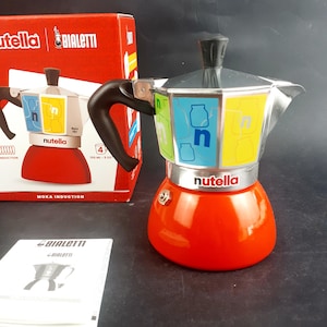 May include: A red and silver Bialetti Moka Induction stovetop espresso maker with a black handle. The coffee maker has colorful panels with the Nutella logo and a capacity of 150 ml (5 oz). The box and instructions are visible in the background.