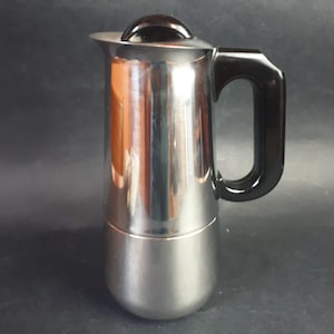 Peut inclure: Une cafetière en acier inoxydable avec une poignée et un couvercle noirs. La cafetière a un corps cylindrique et une base arrondie. La poignée est incurvée et le couvercle a un petit bouton rond. Conçue pour une utilisation sur une cuisinière.