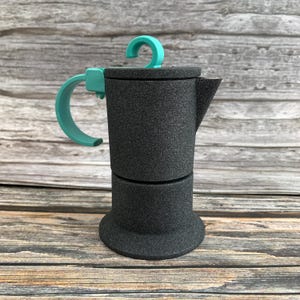 Könnte beinhalten: Ein Espressokocher mit einer texturierten, mattschwarzen Oberfläche und einem leuchtend türkisfarbenen Griff sowie Deckelknopf. Der Espressokocher verfügt über einen zylindrischen Körper, einen breiten Boden und einen markanten Ausguss.