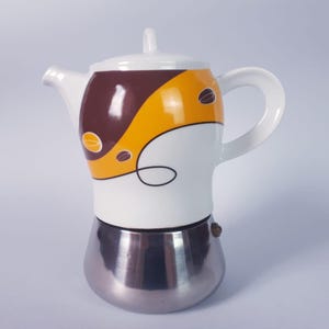 Könnte beinhalten: Weiß-silberne Kaffeemaschine mit braun-gelbem Design. Der Keramikaufsatz hat einen Griff, einen Ausguss und einen Deckel. Das Design umfasst Kaffeebohnen-Illustrationen und eine schwarze Linie. Der Sockel ist silberfarben.