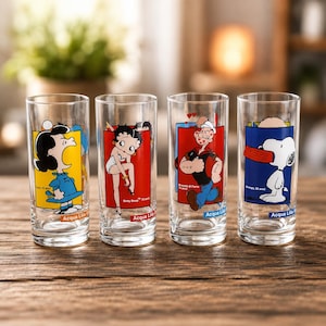 Può includere: Quattro bicchieri trasparenti con disegni di personaggi dei cartoni animati. Ogni bicchiere presenta un personaggio diverso: Lucy dei Peanuts, Betty Boop, Braccio di Ferro e Snoopy. I bicchieri hanno un design rettangolare con uno sfondo colorato e le parole "Acqua Lila" in basso.