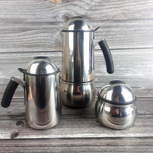 May include: Three stainless steel coffee pots with black handles and lids. The coffee pots are of different sizes and shapes, with a tall, cylindrical pot in the center and a smaller, round pot on the right.