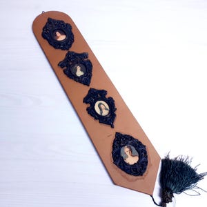 May include: A brown wooden wall hanging with five black ornate frames, each containing a portrait of a woman. The frames are arranged vertically and are connected by a brown fabric strip. A dark green feather tassel hangs from the bottom of the strip.