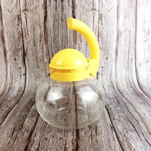 May include: A clear glass water pitcher with a yellow lid and handle. The pitcher has a round body and a curved handle. The lid is also yellow and has a small spout. The pitcher is sitting on a wooden surface.