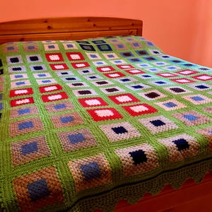 Könnte beinhalten: Eine grüne, blaue, rote und weiße gehäkelte Tagesdecke mit quadratischem Muster. Die Decke liegt auf einem Holzbettgestell.