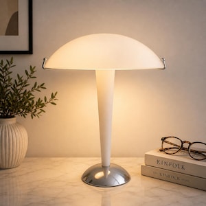 May include: A white table lamp with a mushroom-shaped shade and a chrome base. The lamp has a slender, white stem and a curved, frosted glass shade. The lamp is on a marble surface, next to a vase with greenery and a stack of books.