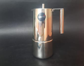 May include: Stainless steel stovetop espresso maker with a cylindrical base and a handle. The coffee maker has a polished finish and a small, round logo that reads "vemi". The handle and top are cylindrical.