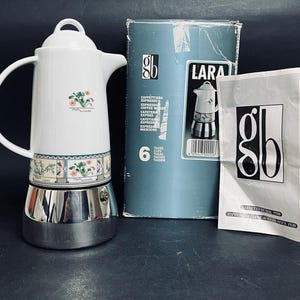 Könnte beinhalten: Eine Espressokanne aus weißer Keramik und silbernem Metall mit einem Blumenmuster am Boden, begleitet von der originalen blauen Pappschachtel mit der Aufschrift Lara und einer weißen Papier-Bedienungsanleitung mit dem gb-Logo.