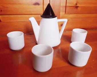 May include: A white ceramic teapot with a black conical lid, a pointed spout, and a square handle, accompanied by three matching white cylindrical cups. The set features a smooth, glossy finish and a clean, geometric design throughout the pieces.
