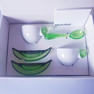 May include: A boxed set of white espresso cups with green handles and matching green saucers. A green spoon is also included. The brand name "guzzini" is visible on a white card inside the box. The set is presented in a white box.
