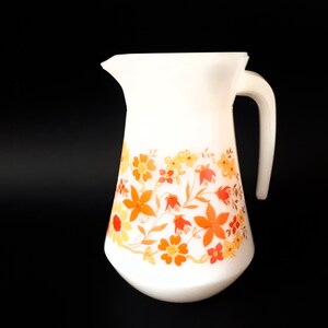 Könnte beinhalten: Weiße Keramikkanne mit einem Blumenmuster in Orange-, Gelb- und Rottönen. Die Kanne hat einen breiten Boden und einen Henkel.