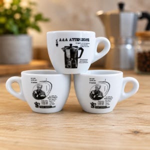 Könnte beinhalten: Drei weiße Keramik-Espressotassen mit schwarzen Aufdrucken. Die obere Tasse zeigt eine Moka-Kannen-Illustration und den Text AAA ATTENZIONE. Die beiden unteren Tassen zeigen ein Porträt eines Mannes, den Text per tutti anche per il nonno und Moka Express.