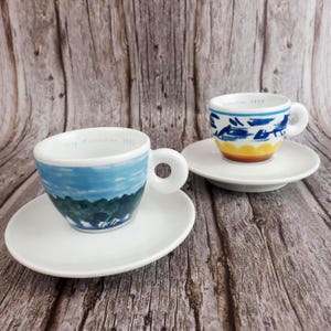 Può includere: Due piccole tazzine da caffè espresso in ceramica bianca con piattini abbinati. Le tazze sono dipinte a mano con disegni di paesaggi in blu, verde e giallo. Il testo "1993 Collection" è stampato sul bordo di ogni tazza. Le tazze hanno un manico rotondo.