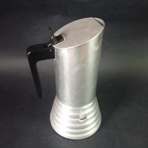 May include: A vintage aluminum stovetop espresso maker with a black handle. The coffee pot has a cylindrical body with a ridged base and a hinged lid. The pot is approximately 8 inches tall.
