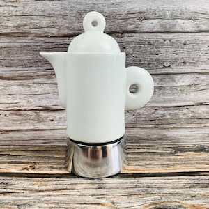 Pode incluir: Uma cafeteira de cerâmica branca com corpo cilíndrico, tampa arredondada com alça circular e uma alça lateral com um orifício. A base da cafeteira é feita de metal polido na cor prata, proporcionando um contraste com a parte superior branca.