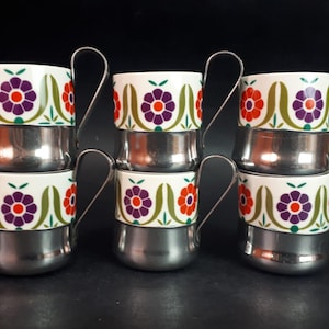 Op de afbeelding: Set van zes vintage koffiekopjes. Elke beker heeft een witte keramische bovenkant met een bloemenontwerp in paars, oranje en groen. De onderste helft is glanzend zilverkleurig metaal. De bekers hebben zilveren metalen handvatten.