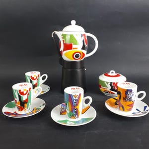 Könnte beinhalten: Ein Pop-Art-Kaffee-Set mit einer schwarzen Espressomaschine, einer Zuckerdose und vier Espressotassen mit Untertassen. Das Set hat ein farbenfrohes, geometrisches Design mit dem Schriftzug "Pop Art" auf den Tassen.