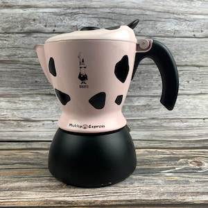 Pode incluir: Uma cafeteira Bialetti Mukka Express com câmara superior rosa claro com manchas pretas de vaca, uma pega preta e uma base inferior preta sólida. O logótipo Bialetti e o texto Mukka Express estão impressos na parte da frente.