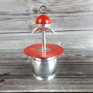 Pode incluir: Uma cafeteira expresso vintage com topo vermelho e corpo prateado. A cafeteira tem um topo redondo vermelho com uma alça de anel prateada e uma haste prateada. A base tem formato cilíndrico prateado. Sobre uma superfície de madeira.