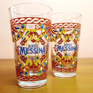 May include: Two clear glass tumblers featuring a colorful Sicilian-inspired pattern in red, yellow, and blue. The glasses display the blue text Birra Messina, the phrase ideata nel 1923, and a 0.2L capacity marking near the top rim of the glass.