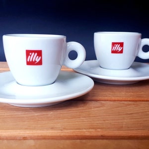 Pode incluir: Duas xícaras de café expresso de cerâmica branca com pires. Cada xícara tem a palavra "illy" impressa em vermelho na frente.