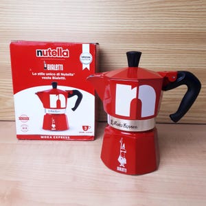 May include: A red Moka Express coffee maker with a black handle and a white "n" on the front. The box next to it is red with the Nutella and Bialetti logos. The text on the box reads "Lo stile unico di Nutella veste Bialetti."