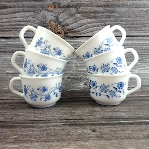 May include: Six white teacups with blue floral patterns are stacked on a wooden surface. Each cup has a curved handle and a delicate design of flowers and vines. The cups are arranged in two stacks of three.