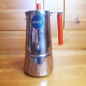 Pode incluir: Uma cafeteira italiana de aço inoxidável com uma alça e parte superior vermelhas. A cafeteira tem um logotipo azul e branco que diz "vemi". A cafeteira está em uma superfície de madeira.
