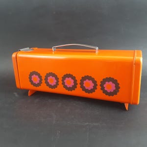 Puede incluir: Una caja metálica naranja con asa y patas plateadas. La caja está decorada con cinco diseños en forma de flor en marrón y rosa. La caja tiene una estética retro y vintage.