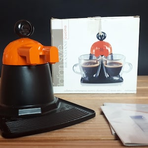 Peut inclure: Une cafetière à espresso noire et orange avec un plateau égouttoir. L'emballage montre la cafetière avec deux tasses de café.