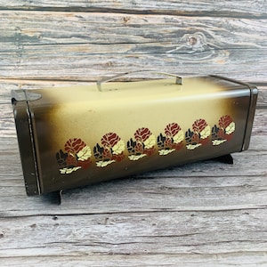 Puede incluir: Una panera rectangular de metal con un acabado degradado en crema y marrón oscuro. La parte frontal está decorada con un patrón repetitivo de motivos florales estilizados en tonos rojos, marrones y amarillos. Incluye un asa de metal plateado.