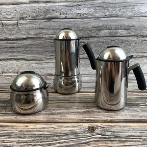May include: A set of three polished stainless steel kitchen vessels consisting of a small rounded sugar bowl with a lid and two taller coffee makers, each featuring a domed lid, a black handle, and a black rim accent on the main body.