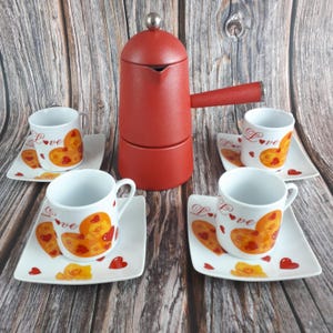 May include: A red stovetop espresso maker with a matching handle and a silver knob, accompanied by four white espresso cups and saucers. The cups and saucers feature a heart design with the word "Love".